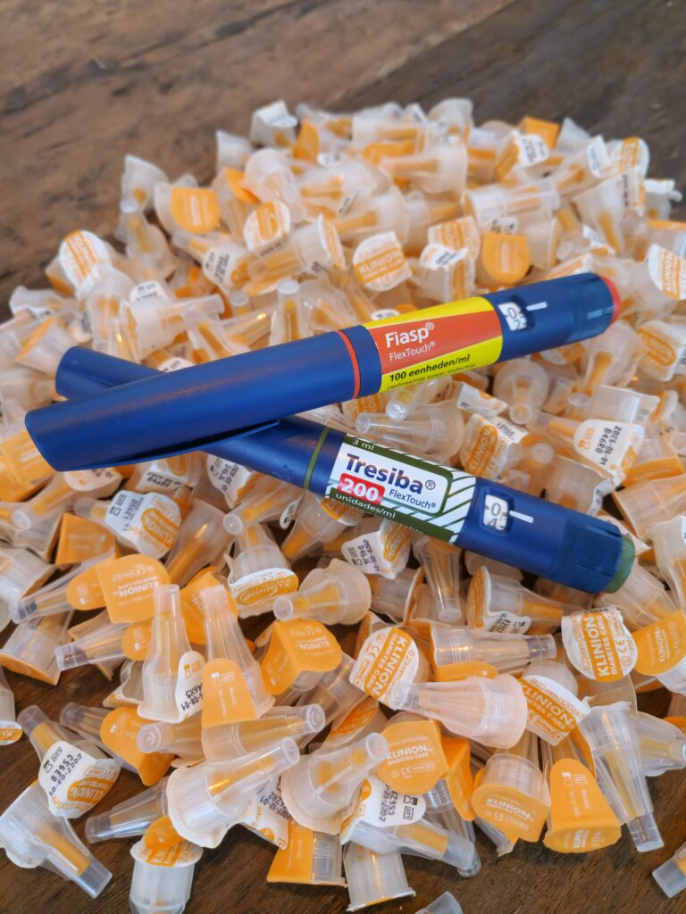 Two blue insulin pens labeled Fiasp and Tresiba lie on top of a large pile of unused orange-and-white insulin pen needles. The image highlights the supplies required for daily diabetes management.