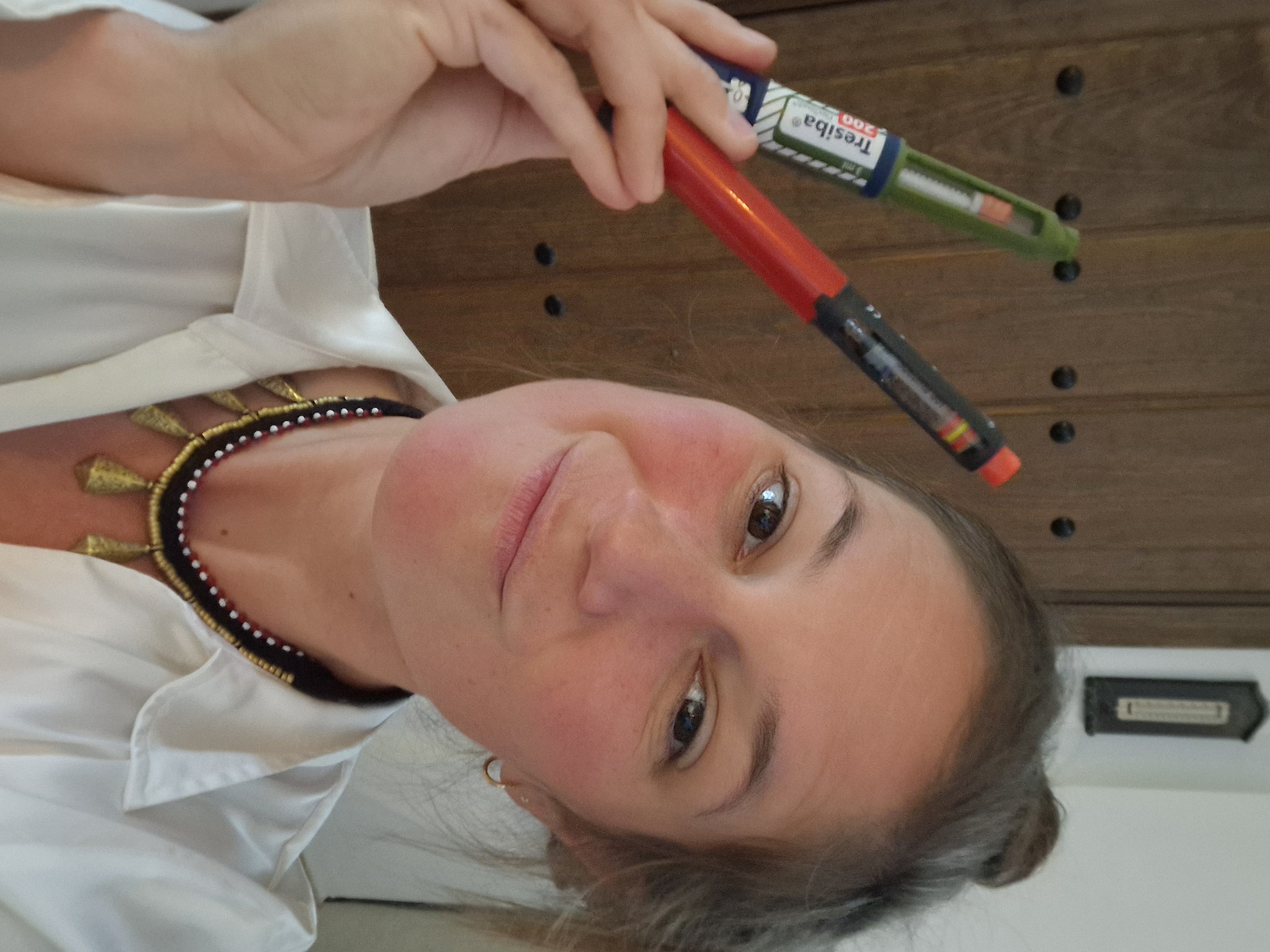 A woman holds up two insulin pens with a confident, slightly playful expression. She is wearing a white shirt and layered necklaces. Behind her is a wooden door. The insulin pens are different colors, showing her daily diabetes management tools.