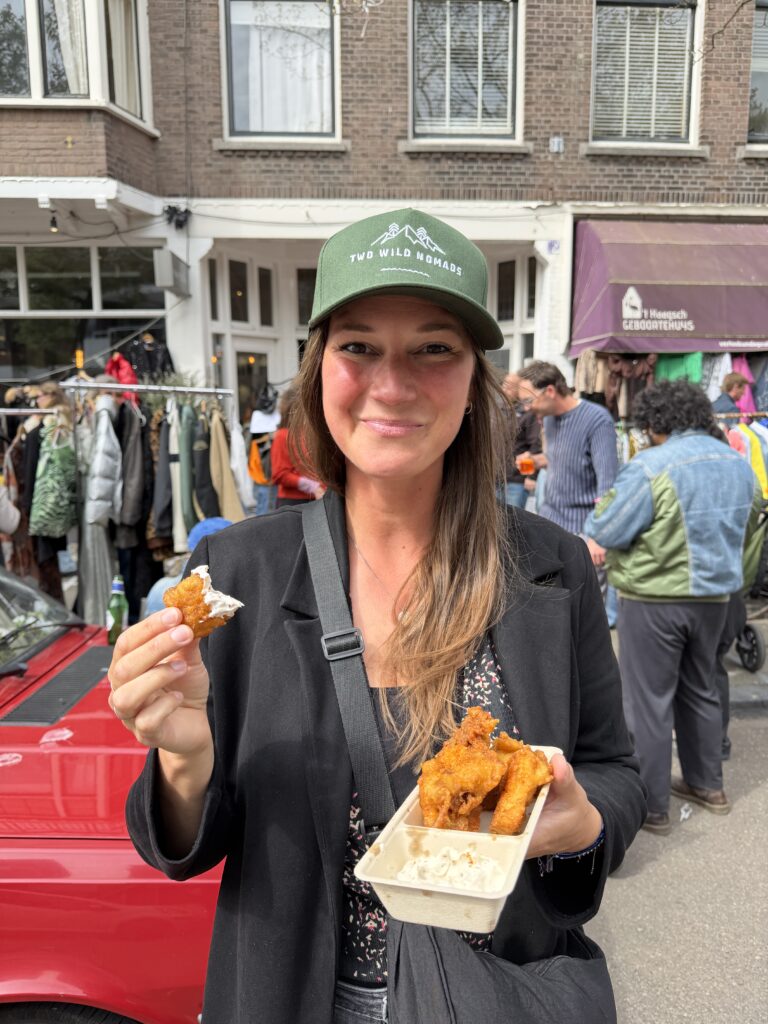 Woman wearing a green Two Wild Nomads cap enjoying street food at a local outdoor market in the Netherlands during full-time world travel.