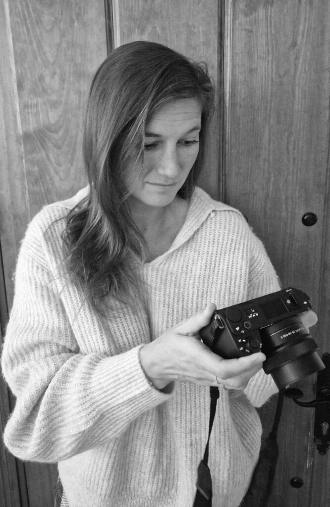 A woman wearing a cozy knitted sweater looks down at a camera in her hands, inspecting the settings. The photo is in black and white, with a wooden door in the background.