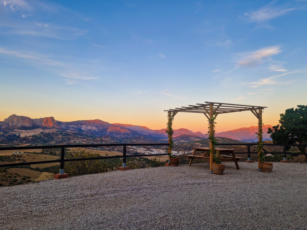 Sunset view from a quiet terrace in rural southern Spain with a wooden pergola, picnic table and potted plants overlooking rolling hills, olive groves and a small white village nestled between warm-colored mountains under a clear evening sky.