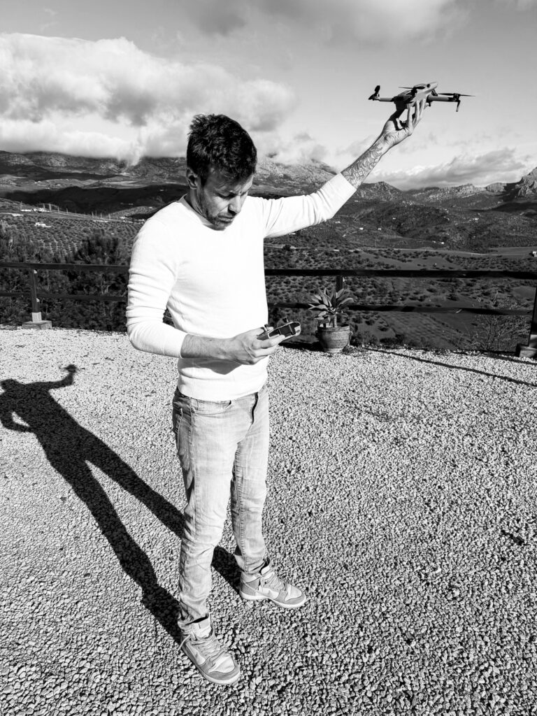 A man stands outdoors on a gravel terrace holding a drone up in one hand while looking down at a controller in his other hand. The scene is in black and white, with mountains and clouds visible in the background.