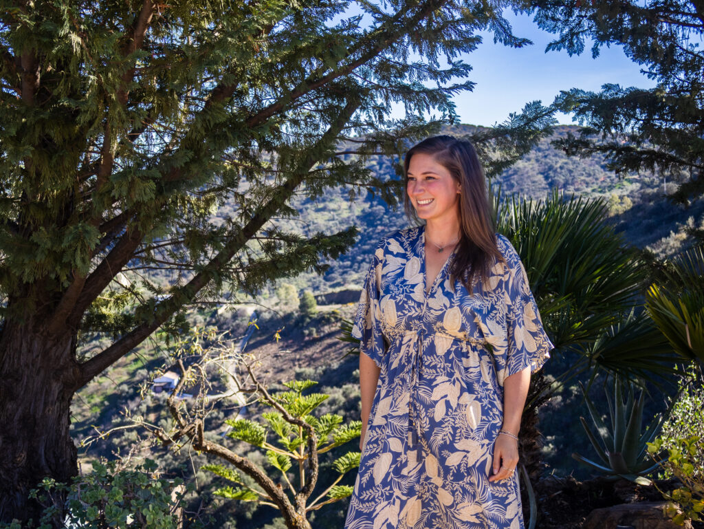 A smiling woman stands outdoors under a large tree, wearing a blue and beige floral dress. Behind her, sunlit hills and green plants create a peaceful Mediterranean landscape.