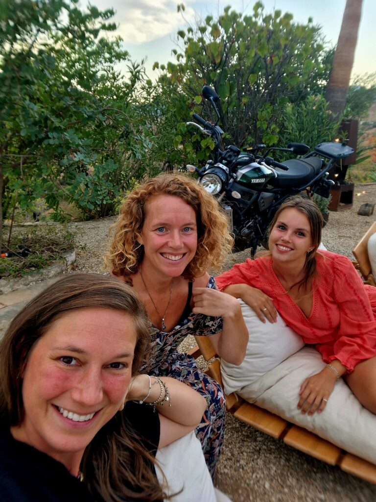Three women smiling together outdoors in southern Spain with a motorcycle behind them, enjoying a relaxed evening surrounded by nature.
